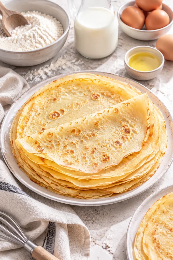 Dünne Pfannkuchen auf einem weißen Teller mit neutralem Öl und Holzlöffel